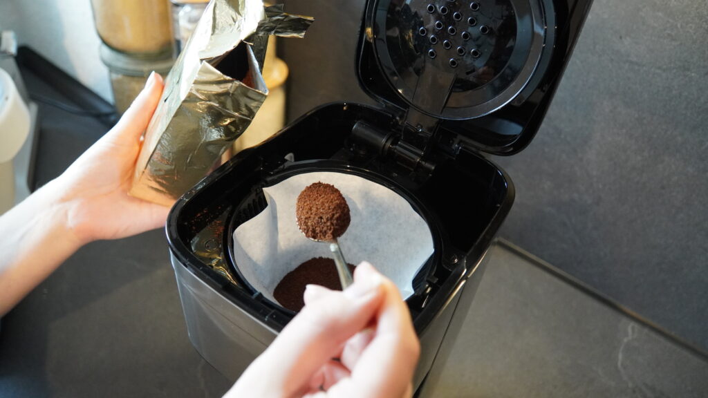 Eine Person hält einen Löffel mit Kaffeepulver über einen Filter in einer schwarzen Kaffeemaschine Let’s Brew Elegant.