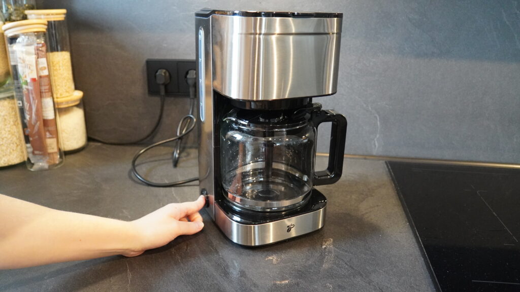 Kaffeemaschine Let’s Brew Elegant  mit Glaskanne und einem Bedienknopf auf der Vorderseite, auf einer dunklen Arbeitsfläche platziert.